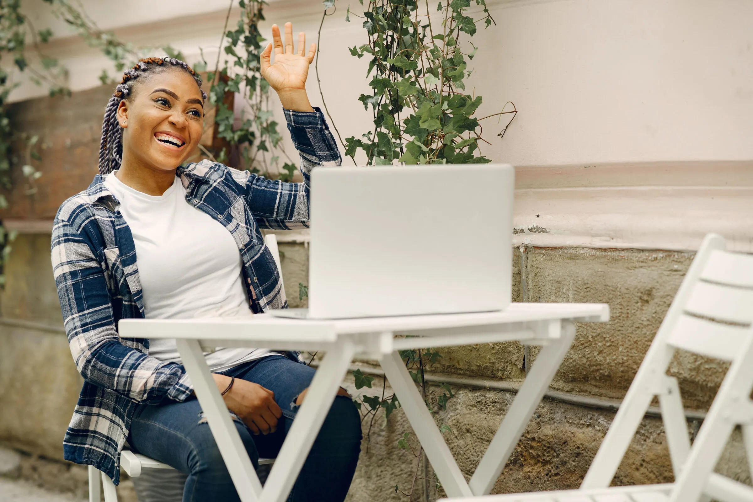 A smiling young woman waves from her laptop at an outdoor table